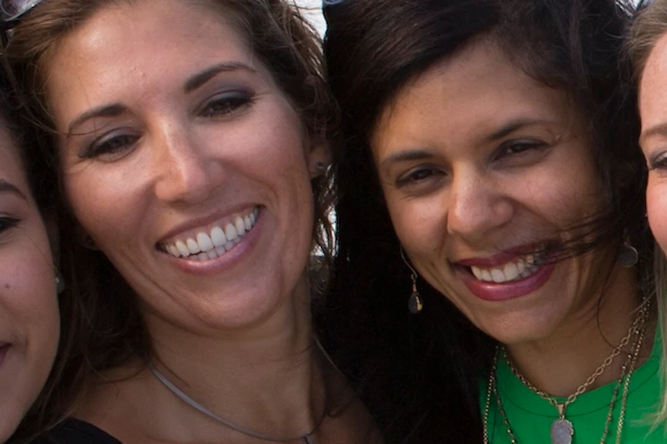 Four women taking a selfie in front of a waterfall, smiling. The sky is clear with some white clouds.