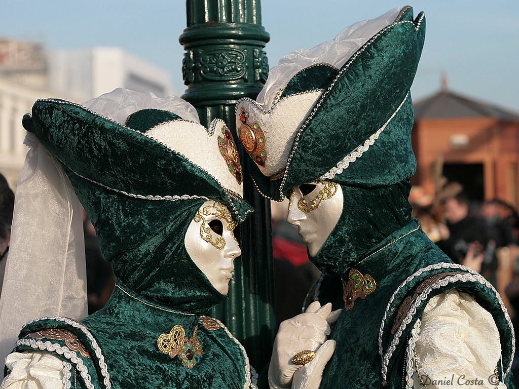 Zwei Personen in grün-weißen venezianischen Karnevalskostümen und Masken stehen dicht beieinander in der Nähe einer geschmückten Stange.