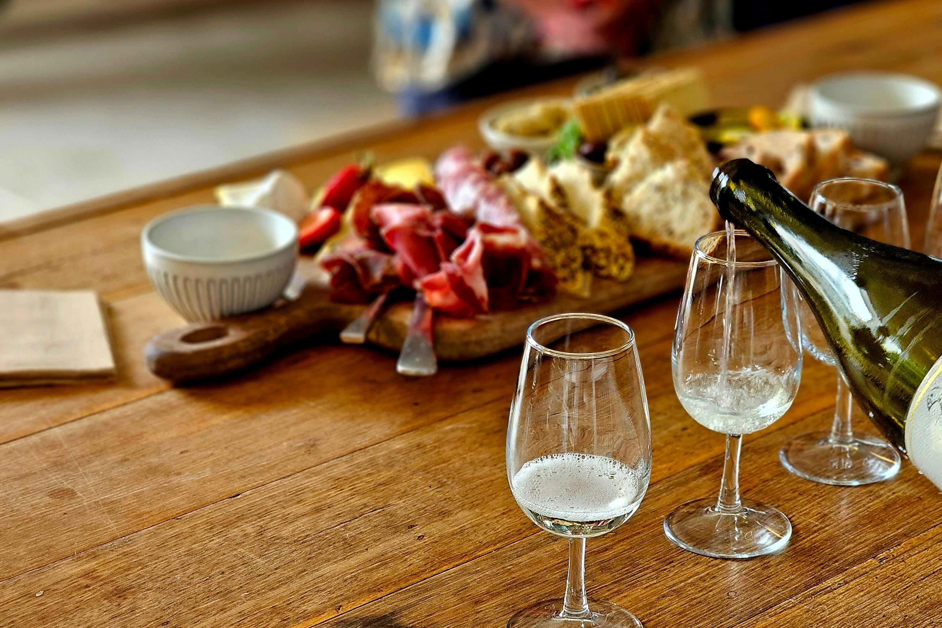 A wooden table with a charcuterie board of meats, cheeses, and bread, with two wine glasses being filled with white wine.