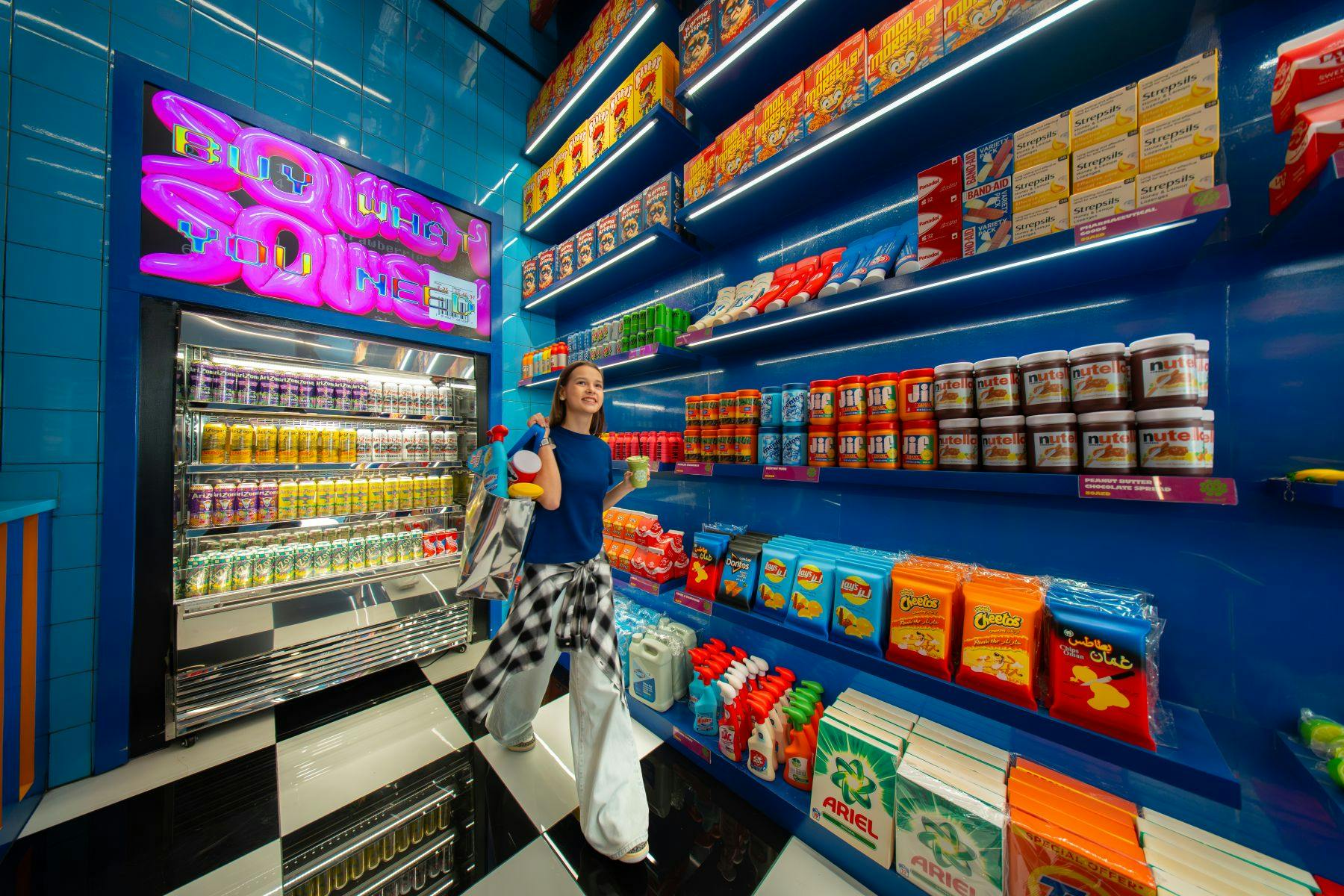 Eine Person betritt ein hell erleuchtetes Geschäft mit bunten Regalen und einer Leuchtreklame, in der Hand eine Einkaufstasche und ein Getränk.