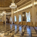 A grand, ornate hall with marble floors, chandeliers, draped windows, and tall columns. No people are present.