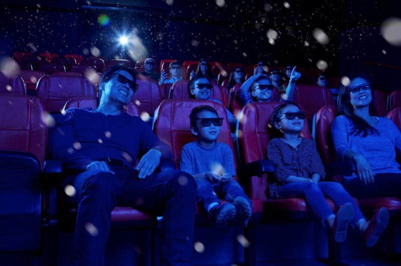 Persone con occhiali 3D sedute in un cinema con poltrone rosse che guardano un film con effetti speciali visibili in una stanza buia.