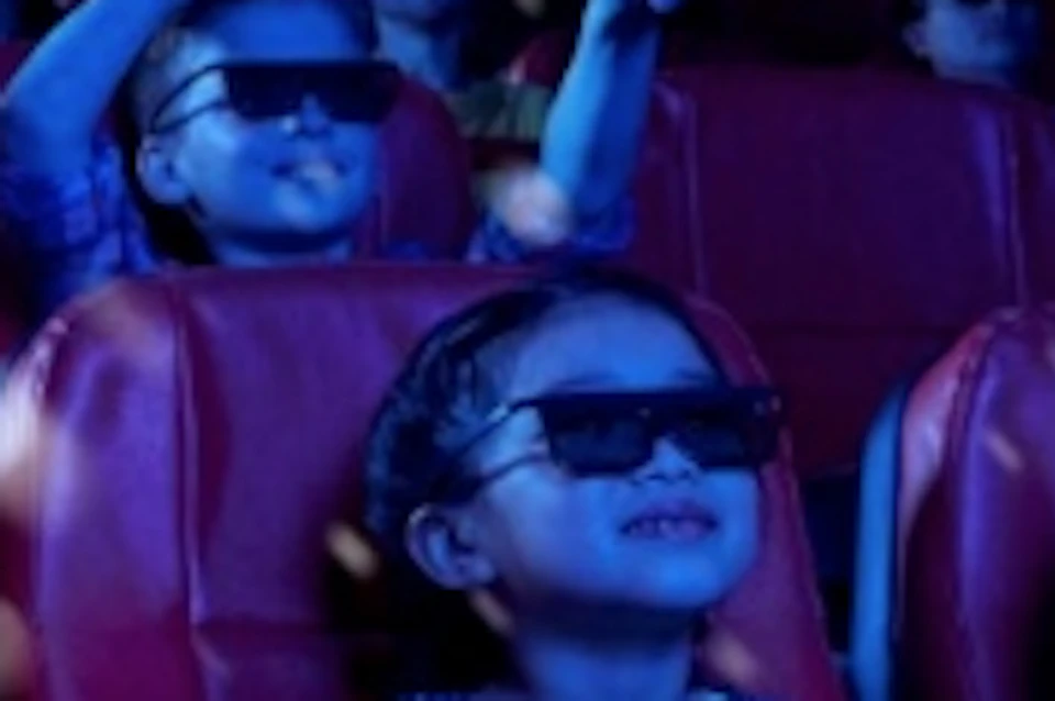 People wearing 3D glasses seated in a cinema with red seats, watching a film with visible special effects in a dark room.