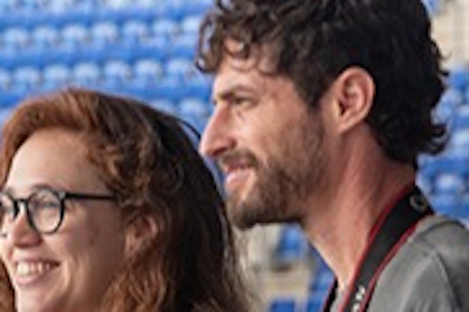 Four people standing in the seating area of a stadium, looking out towards the field, with rows of blue seats around them.