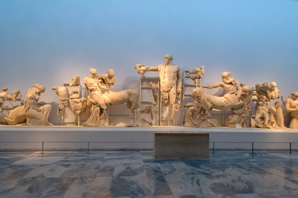 A collection of ancient marble statues depicting human and animal figures displayed in a museum against a blue wall.