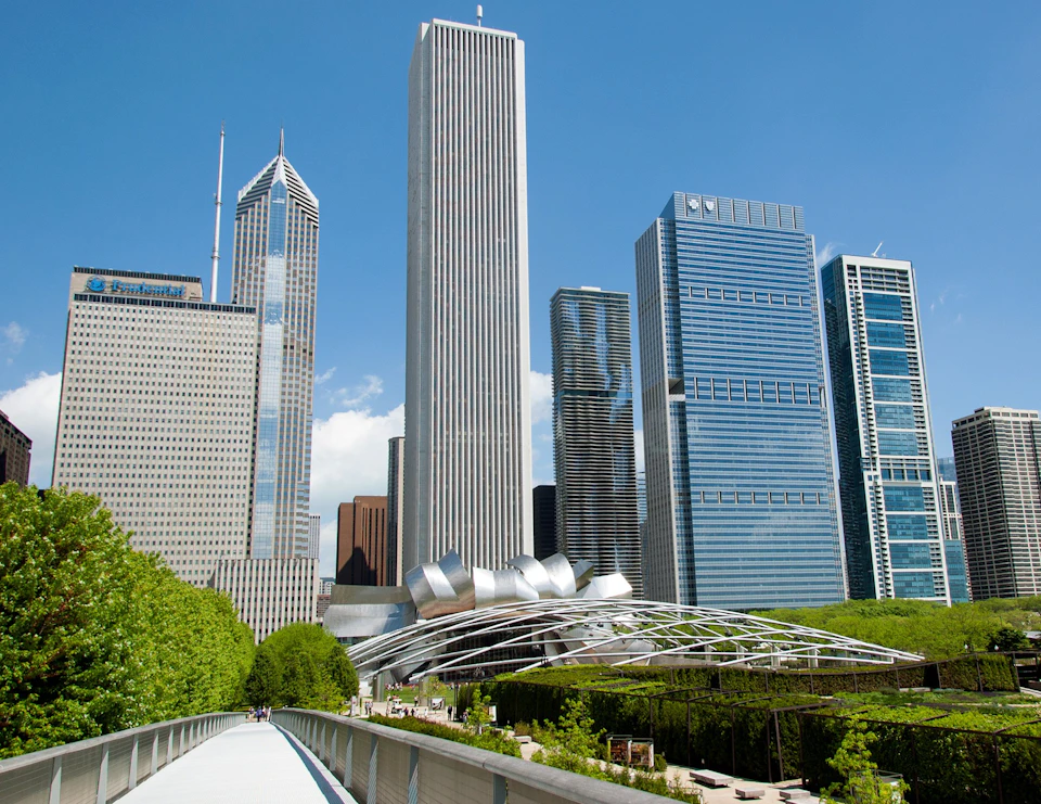 Modern skyscrapers and a metallic, sculptural structure surrounded by greenery, viewed from an elevated walkway under a clear blue sky.