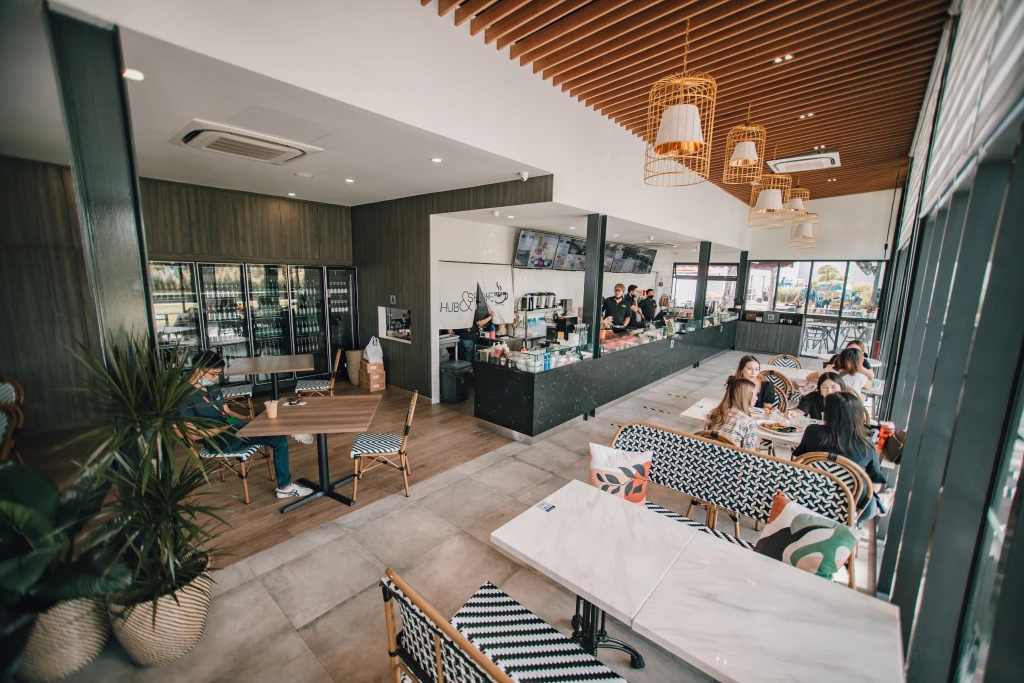 A modern cafe with wooden and white decor, a coffee counter, tables, and people sitting and talking.