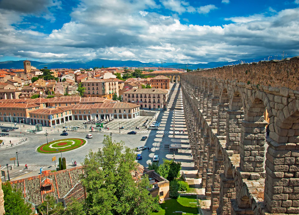 A Roman aqueduct stretches across a historic city with red-tiled roofs, a central plaza, and a mountainous background under a blue sky.