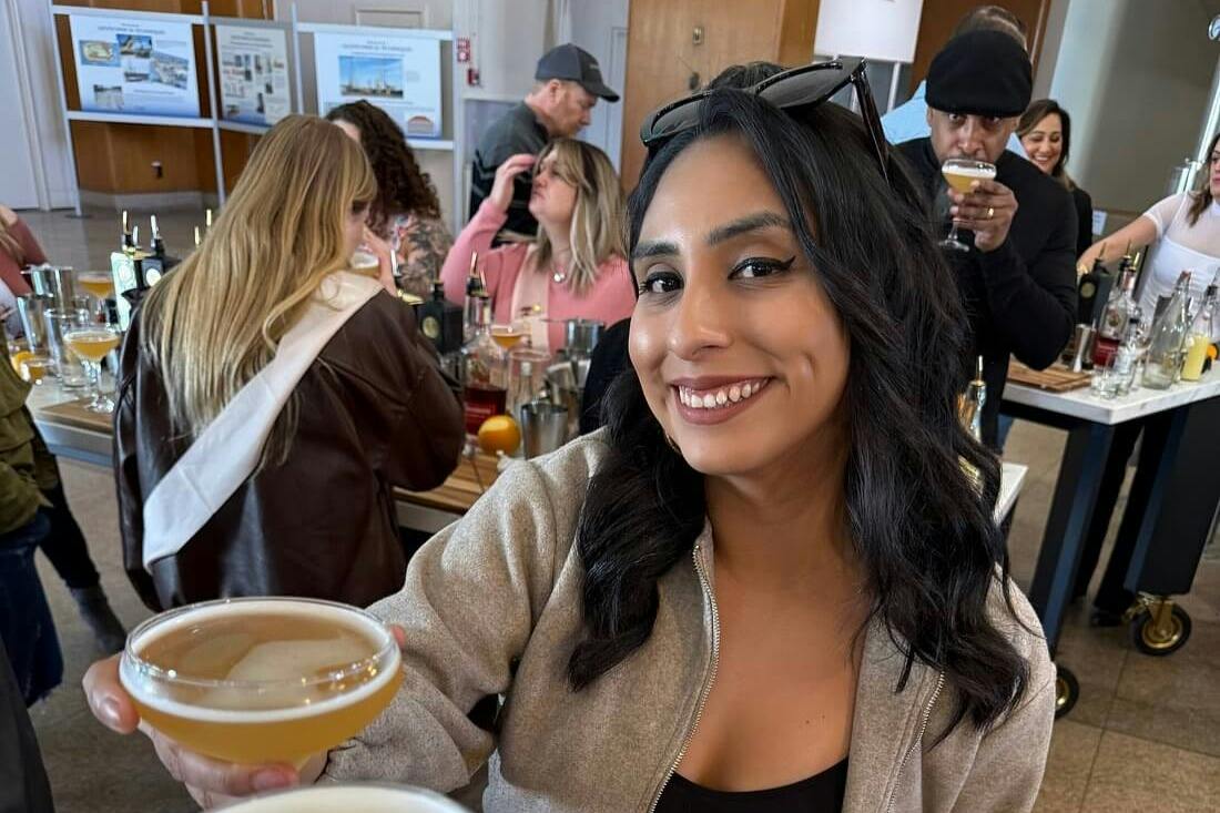 A woman smiling at the camera in a busy indoor setting, with people in the background, some holding drinks.