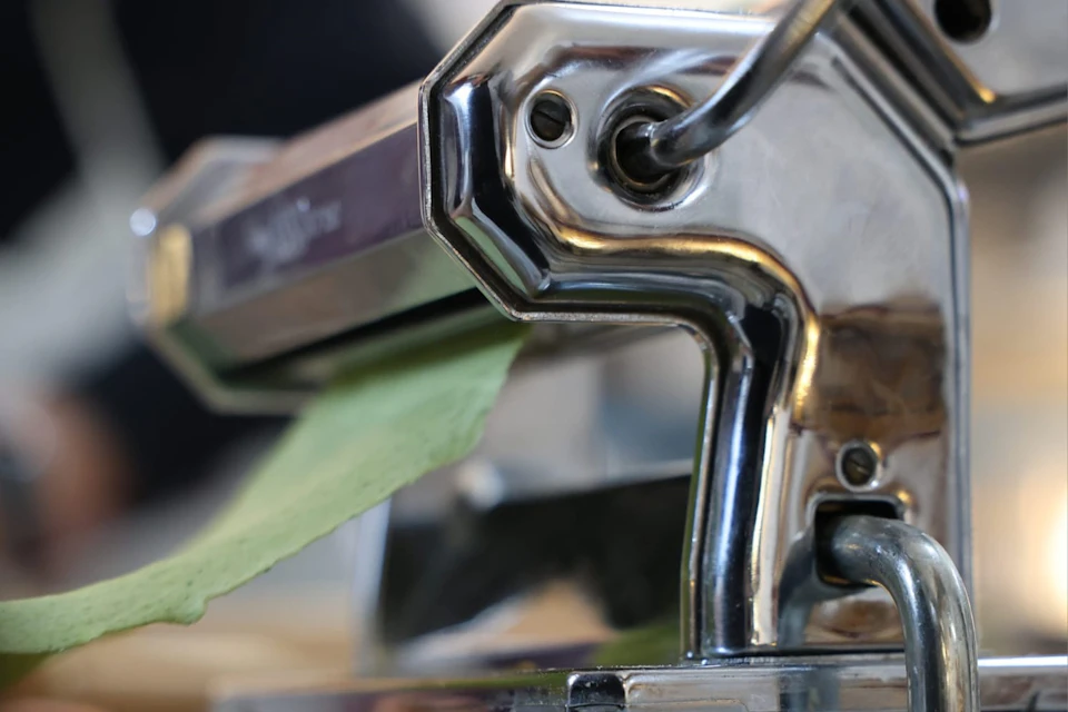 A close-up of a metal pasta maker with green pasta dough being rolled through it.