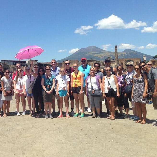 一群人站在户外，后面有废墟和一座山。 一个人拿着一把粉红色的雨伞。