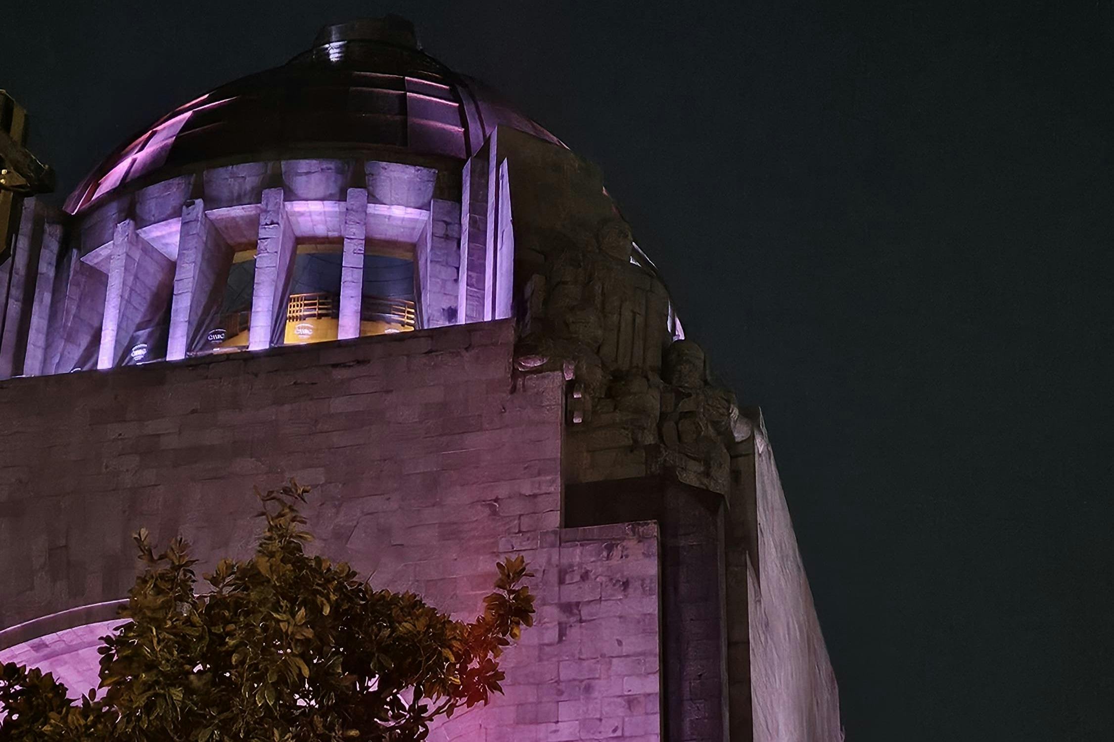 Een stenen gebouw met een koepel verlicht door paarse lichten bij nacht, met een deel van een boom zichtbaar op de voorgrond.