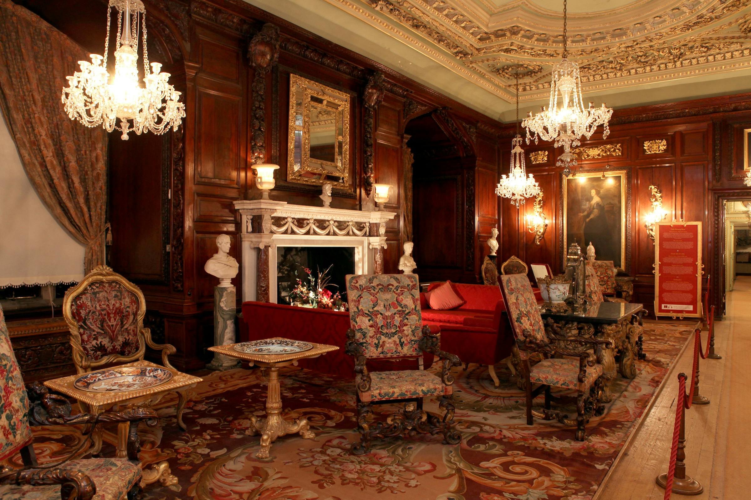 Ornate room with a decorated fireplace, vintage furniture, chandeliers, and statues. Detailed wood paneling and a patterned carpet.
