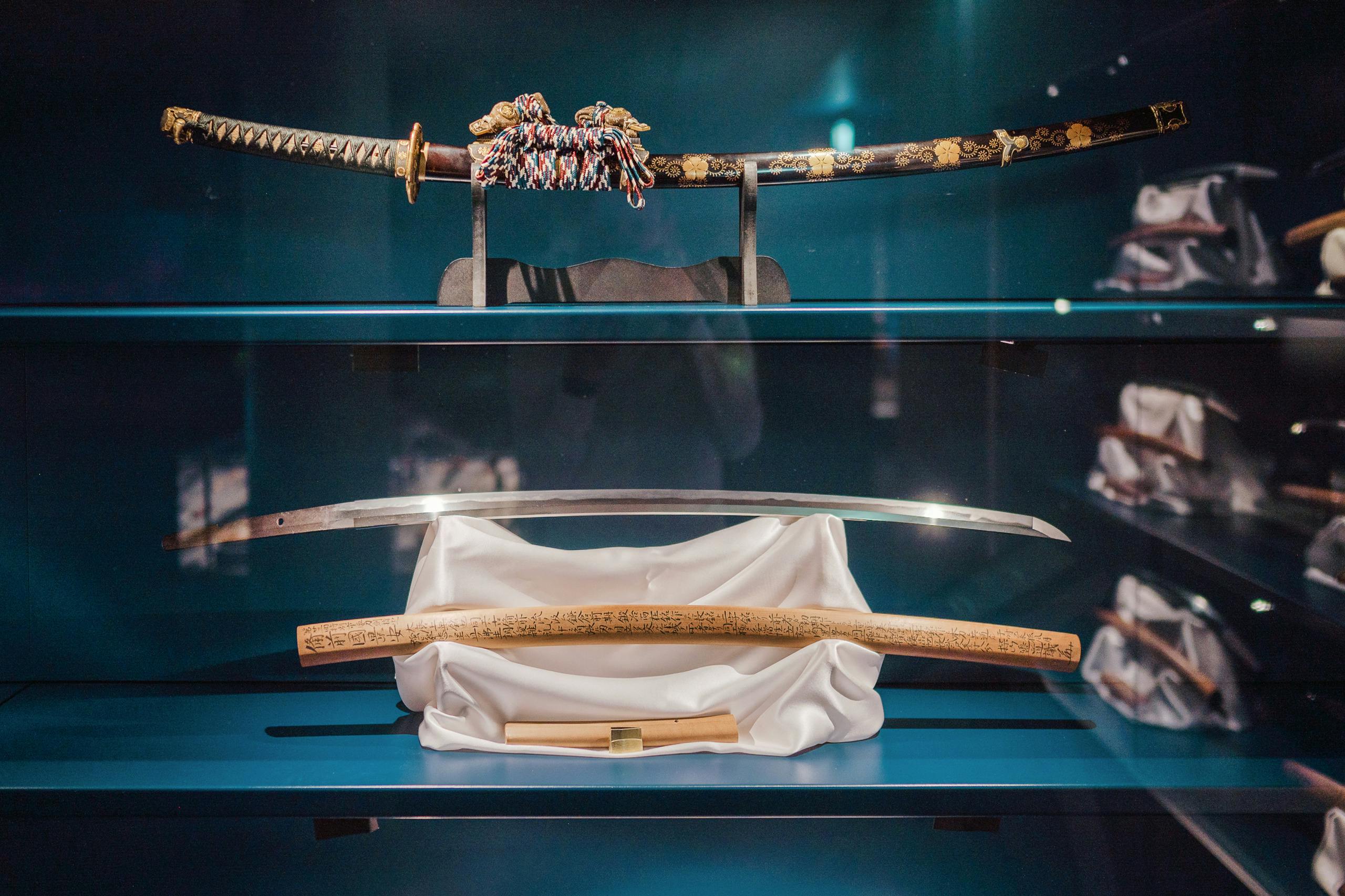 A display case with two Japanese swords: one sheathed with ornate decorations and one unsheathed on a white cloth.