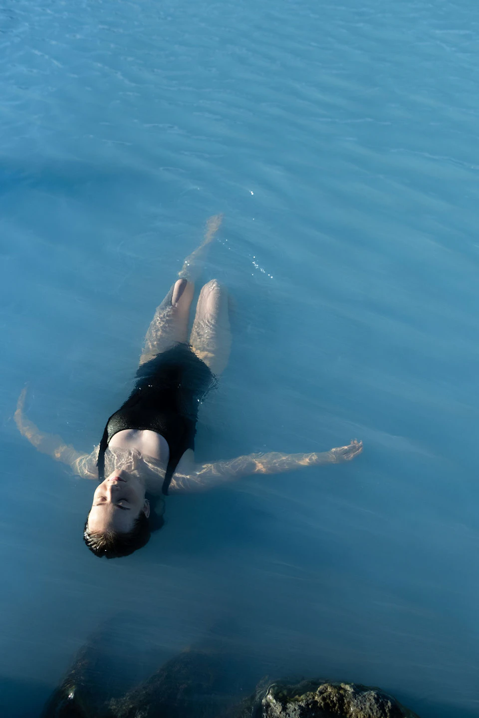A person in a black swimsuit floating on their back in calm blue water under sunlight.