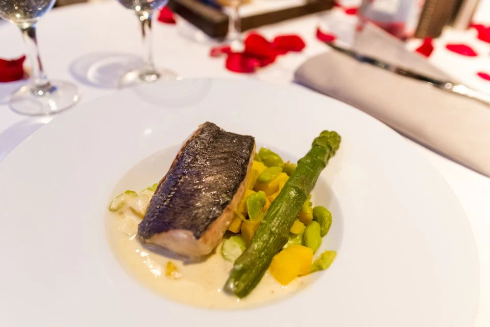 A plate of fish with asparagus and vegetables, served on a white tablecloth with rose petals and glassware in the background.
