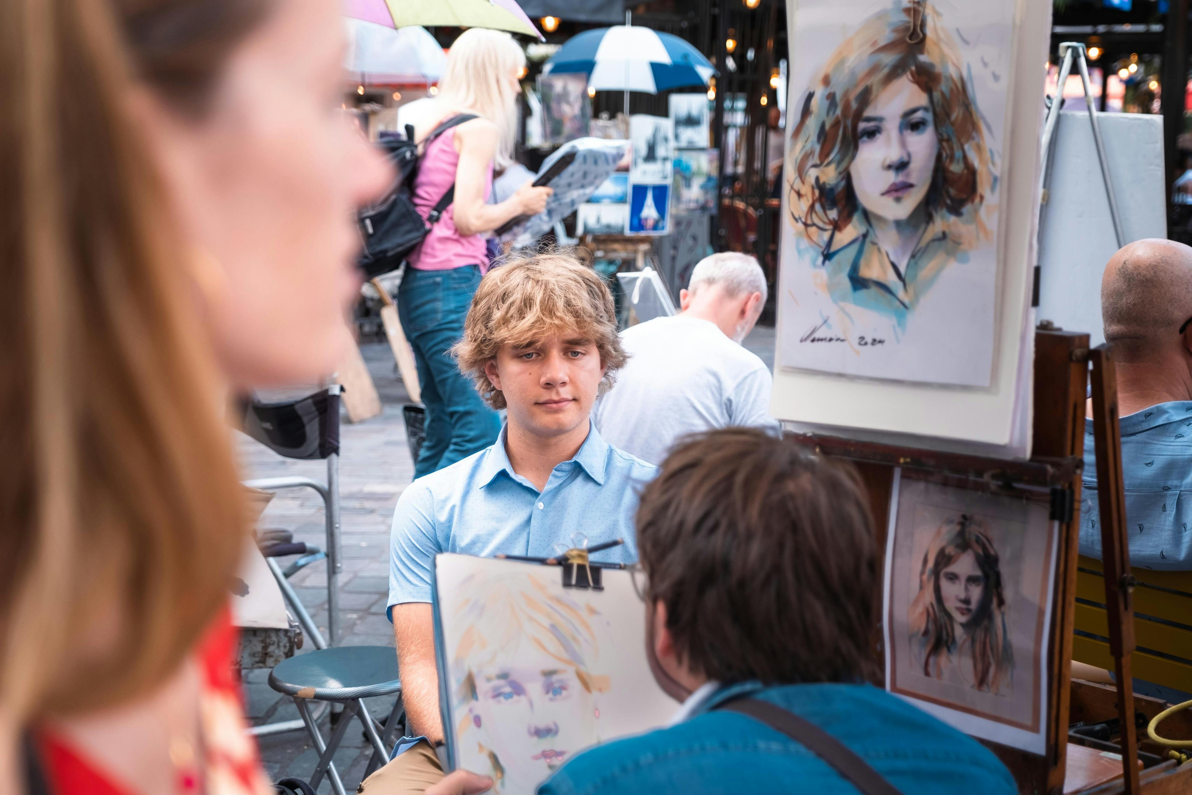 An artist sketches a young man in a busy outdoor setting, with other people and artwork visible in the background.