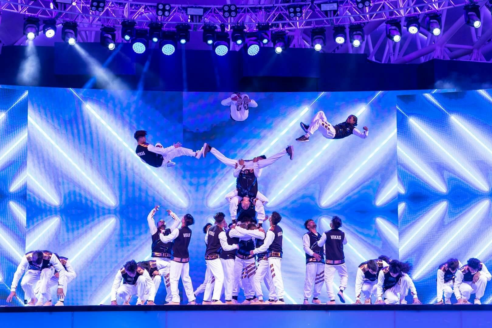Taneční štáb vystupuje na jevišti s dynamickým modrým osvětlením se synchronizovanými salty a akrobatickými kaskadérskými kousky.