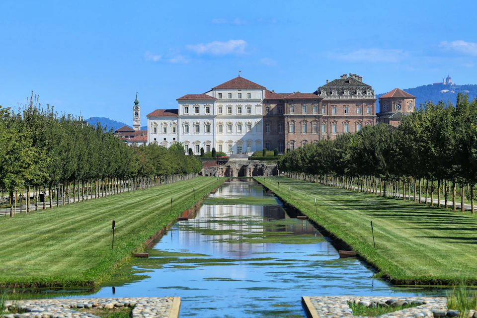 A stately building with a red roof surrounded by a landscaped garden, with a long reflective waterway in the foreground.