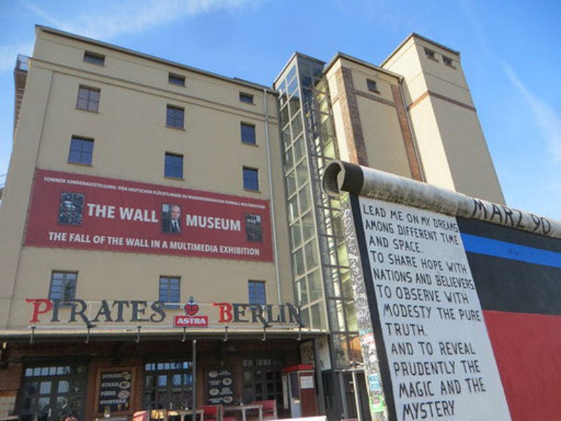 Facciata di un edificio con striscione "The Wall Museum" e parte del Muro di Berlino con testo. Il cartello recita "PIRATI BERLINO".