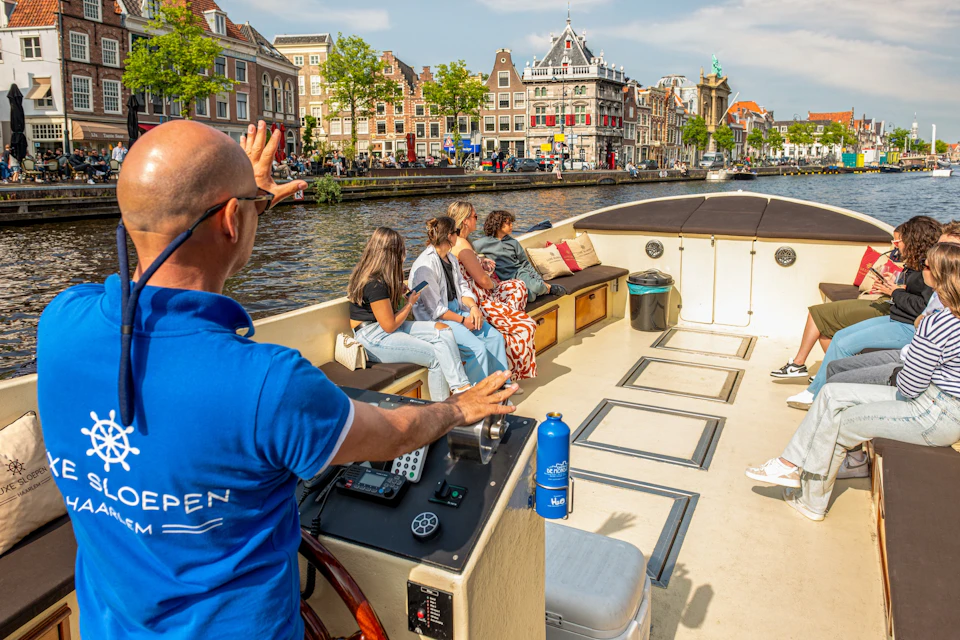 People seated on a boat in a canal, with historic buildings and trees along the water in a sunny urban setting.