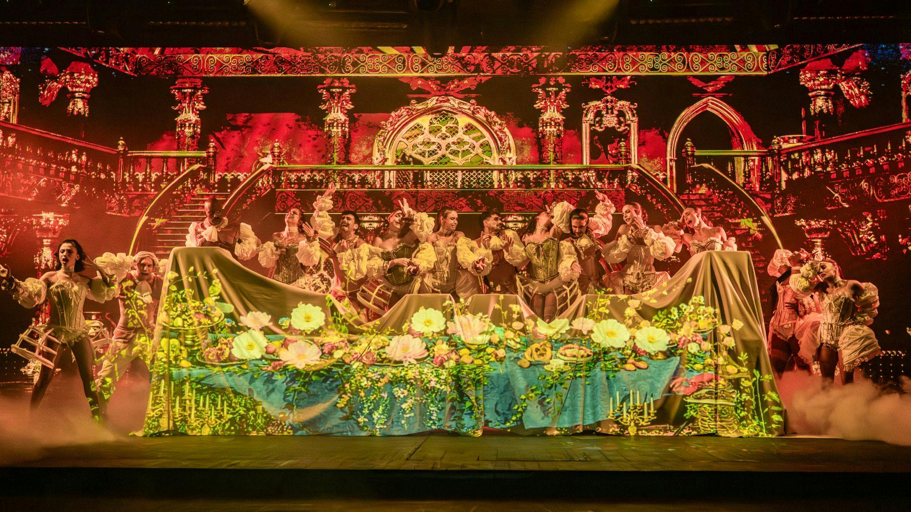 Um grupo de artistas em trajes ornamentados no palco com uma vibrante iluminação de fundo floral e decorativa.
