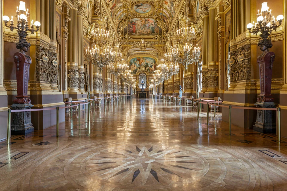 A grand hall with ornate gold decorations, chandeliers, and a polished floor, featuring a central design and a statue in the distance.