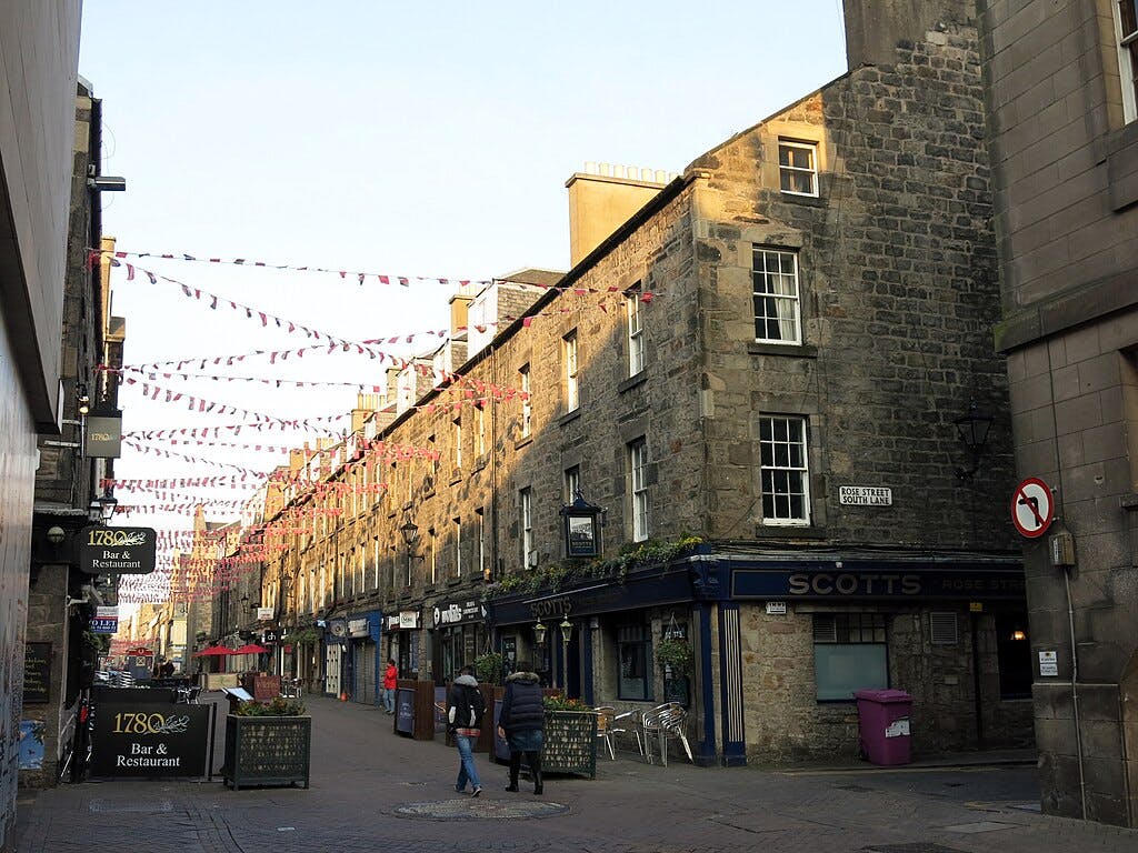 A narrow cobbled street lined with old stone buildings, hanging bunting, and people walking. Signs for bars and restaurants are visible.