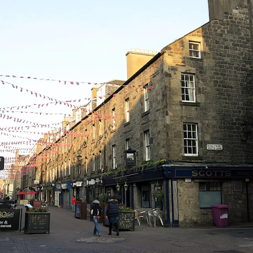 En smal kullerstensgata kantad av gamla stenbyggnader, hängande vimplar och människor som går. Skyltar för barer och restauranger är synliga.