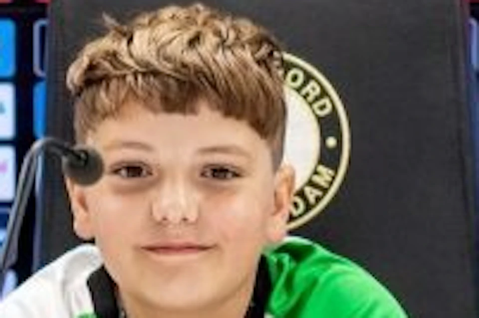 Two young boys sitting at a press conference table in front of a sponsor backdrop, appearing at a sports-related event.