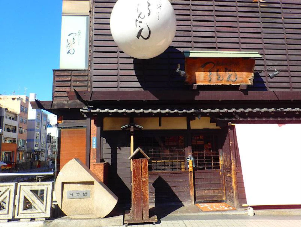 A traditional Japanese storefront with wooden accents, signage, a large round lantern, and a decorative stone monument.