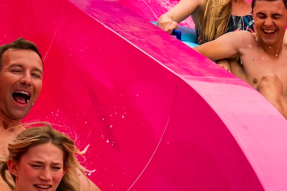 Four people on blue inflatables slide down a bright pink water slide, surrounded by twisting yellow and green slides.