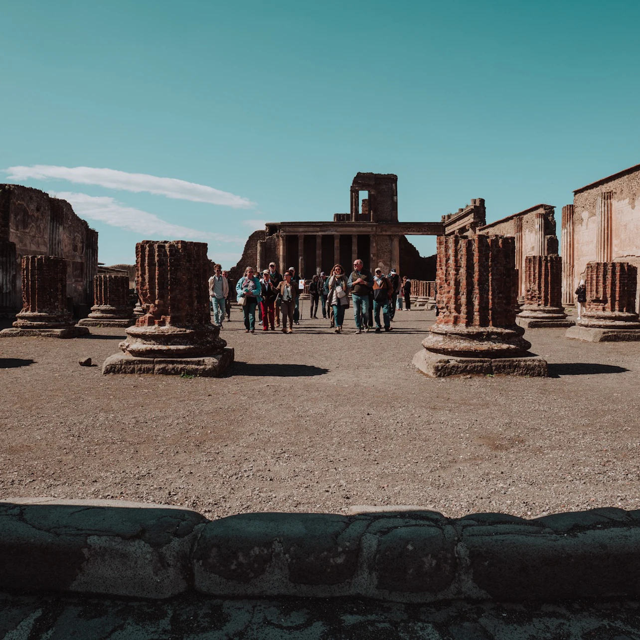 A group of tourists explore ancient ruins with large stone columns under a clear blue sky.