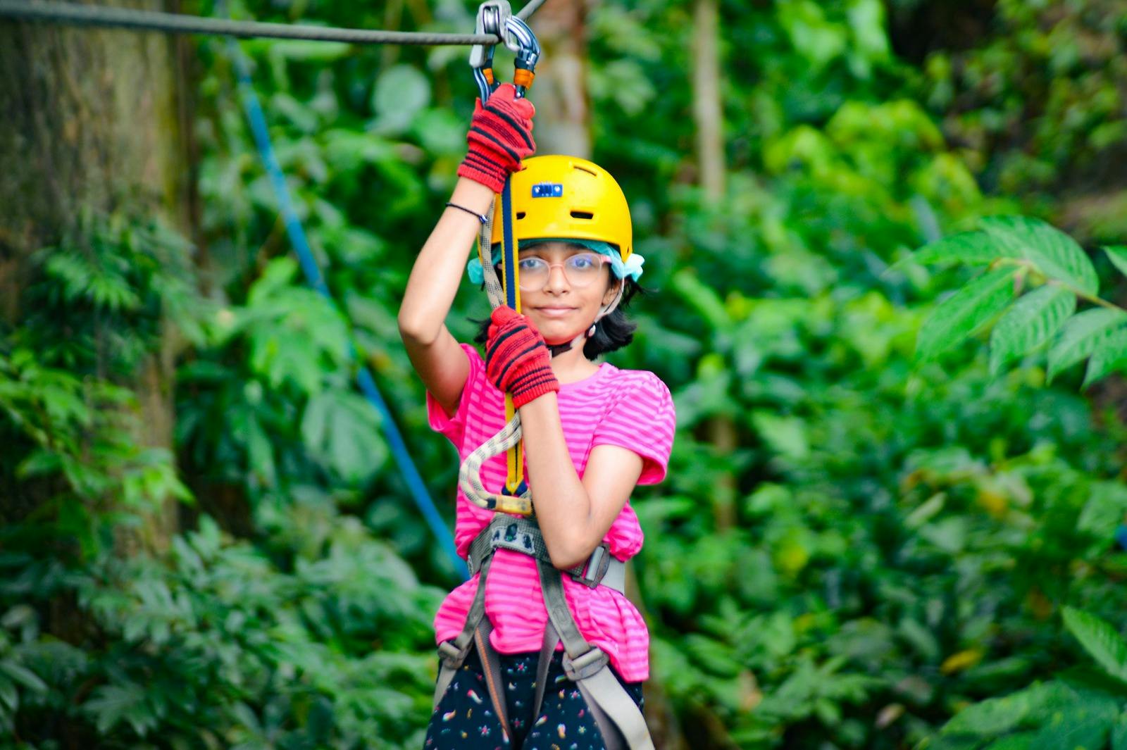 Un niño con casco amarillo y camiseta rosa se sujeta a un arnés de tirolina en un frondoso bosque verde.