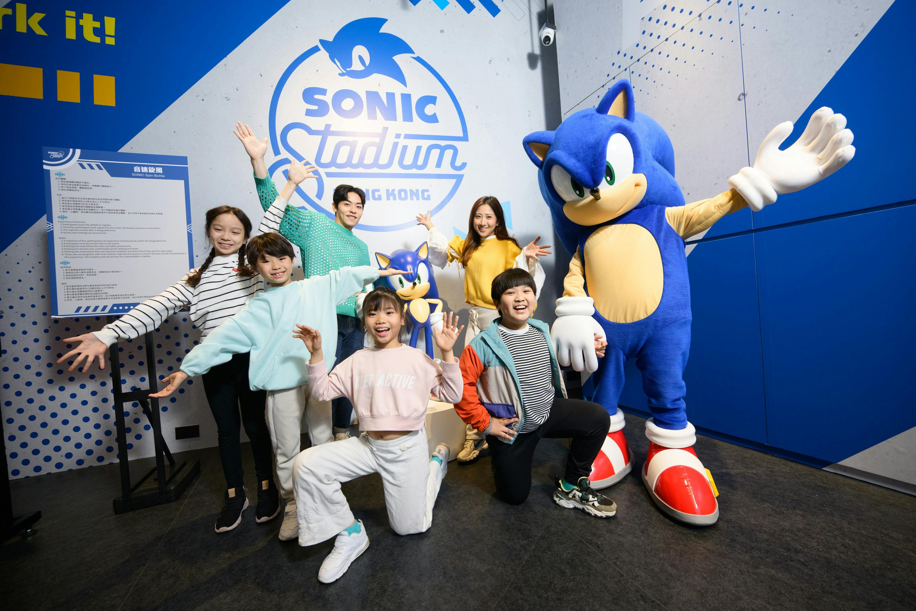 A group of children and adults posing enthusiastically with a Sonic the Hedgehog statue in front of a Sonic-themed backdrop.