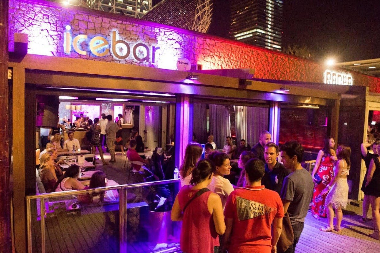 Des gens socialisent à l'extérieur d'un bar éclairé la nuit, avec des lumières colorées et des bâtiments urbains en arrière-plan.