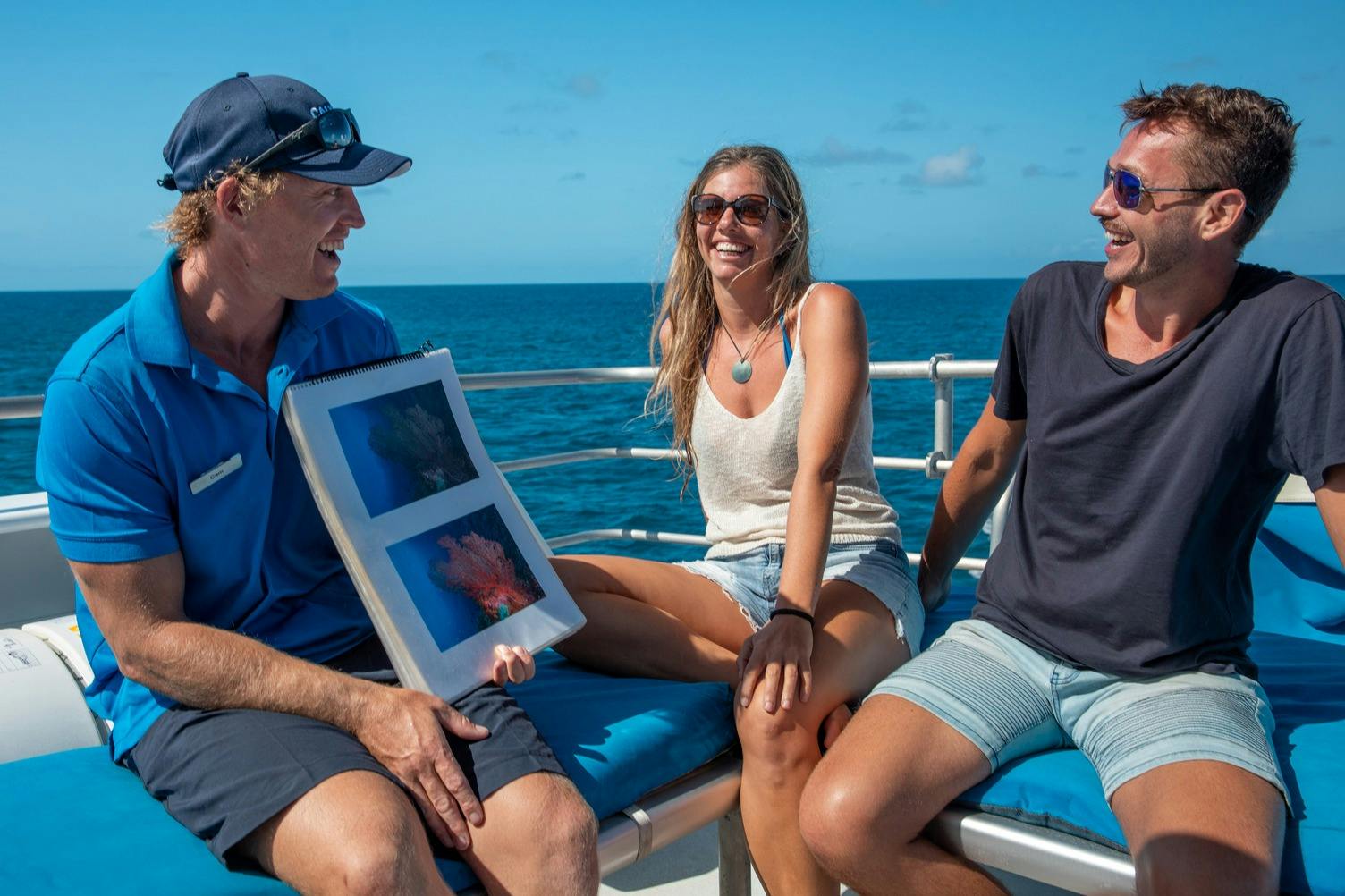 Tres persones en un vaixell somrient i sostenint un llibret amb imatges submarines, amb l'oceà de fons.