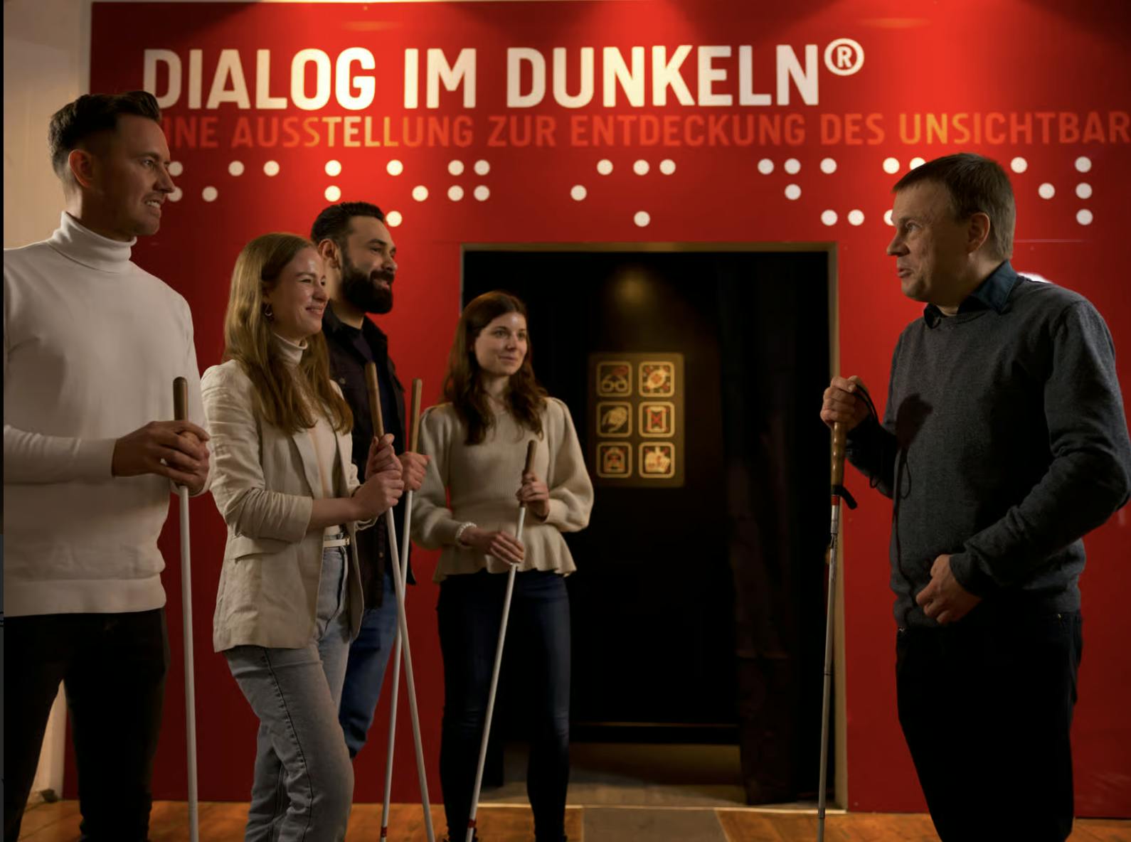 五个人拿着白色手杖，站在一堵红色的墙前，上面写着“Dialog im Dunkeln”的文字和盲文符号。