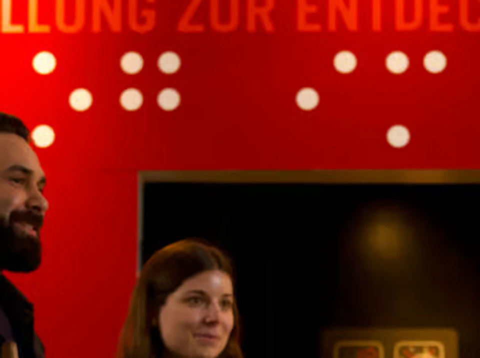 Five people hold white canes and stand in front of a red wall with "Dialog im Dunkeln" text and Braille symbols.