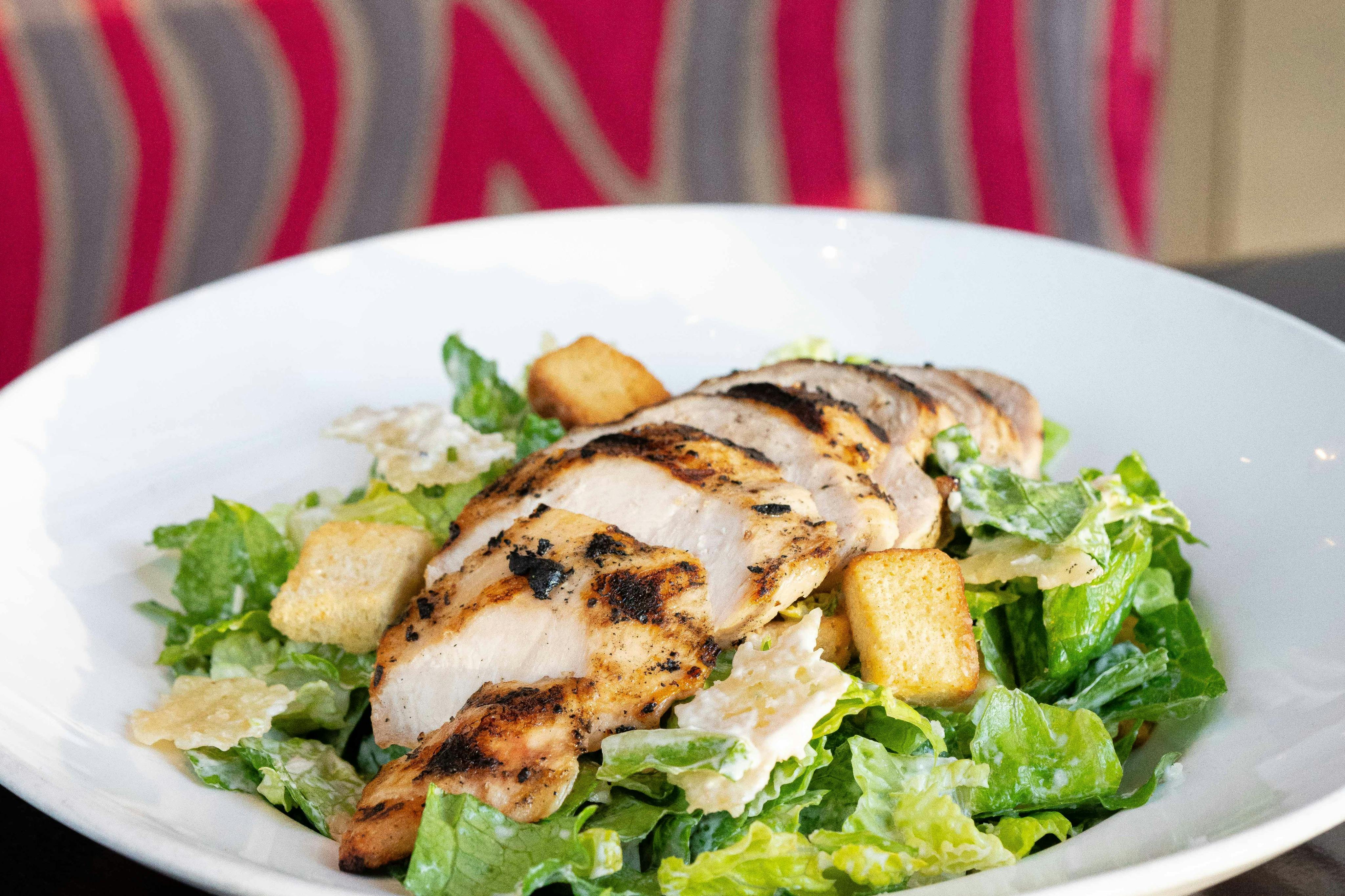 A Caesar salad topped with sliced grilled chicken and croutons, served in a white bowl with a colorful background.