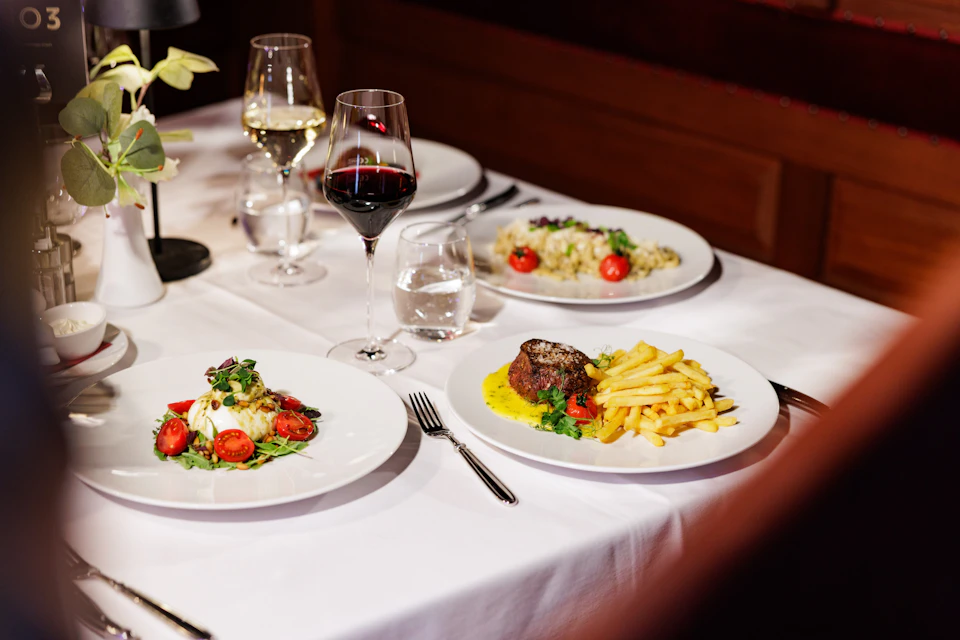 A table set for a meal with three plates of gourmet food, wine glasses, and a water glass on a white tablecloth.