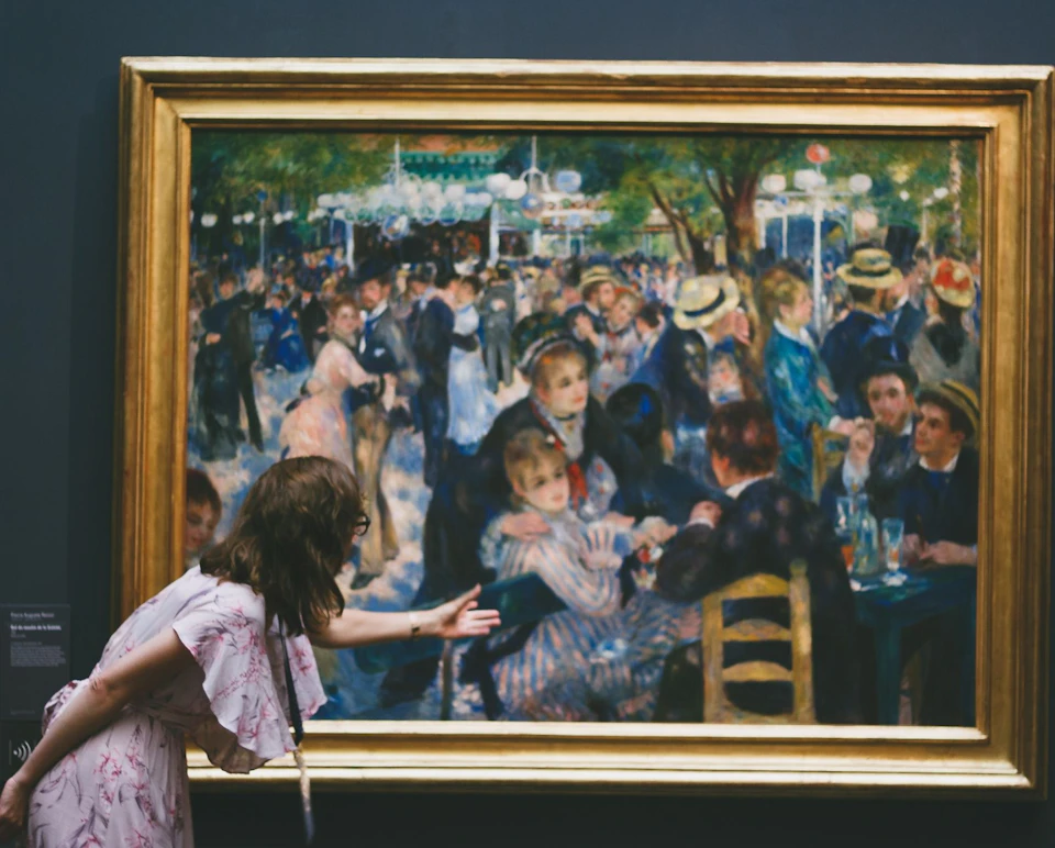 A woman leans towards a large framed painting depicting a lively outdoor social gathering with many people in period clothing.