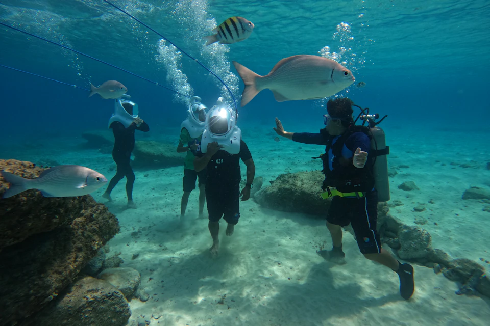 Group of people underwater wearing helmets, surrounded by fish and a scuba diver giving instructions. Clear blue water, rocky seabed.