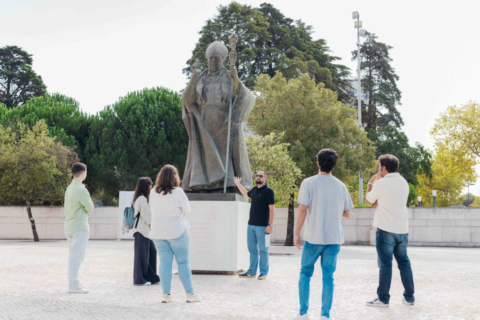 A group of people gather around a statue of a figure holding a staff, with trees in the background.