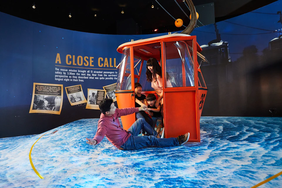 A family interacts with a red rescue capsule exhibit over a simulated ocean surface, with informational text in the background.