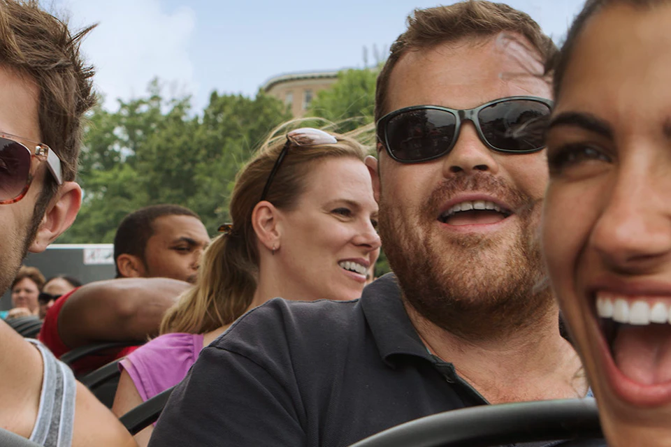 A group of smiling adults wearing sunglasses and casual clothes, enjoying a ride on a bus or shuttle with trees in the background.