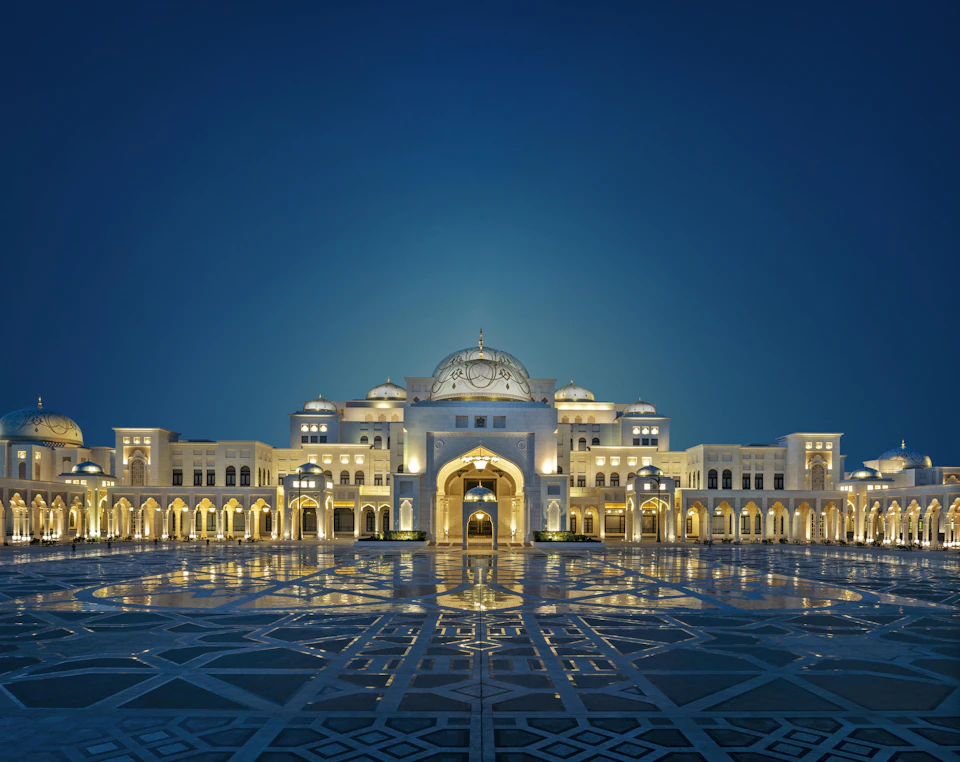 An illuminated grand palace with multiple arches and a central dome, reflected on the ornate pavement under a deep blue sky.