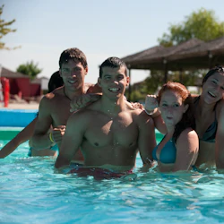 En grupp på sex leende unga vuxna står i en pool, med träd och skuggade strukturer i bakgrunden.