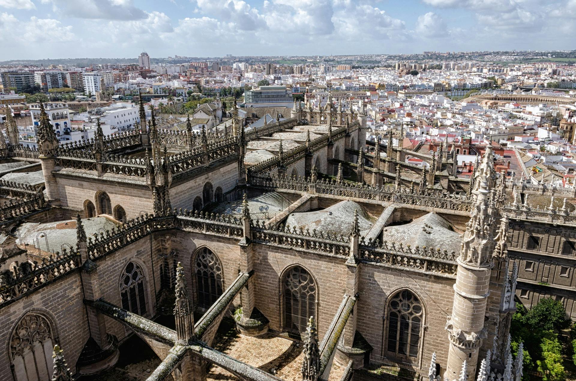 Les toits des cathédrales gothiques aux flèches complexes surplombent un paysage urbain densément peuplé sous un ciel partiellement nuageux.