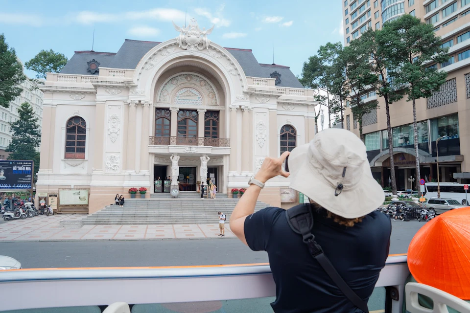 A person wearing a hat and backpack takes a photo of an ornate building with an arched entrance and detailed facade.